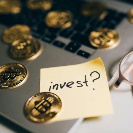 A close-up of bitcoin coins on a laptop with eyeglasses and a sticky note reading 'invest?'