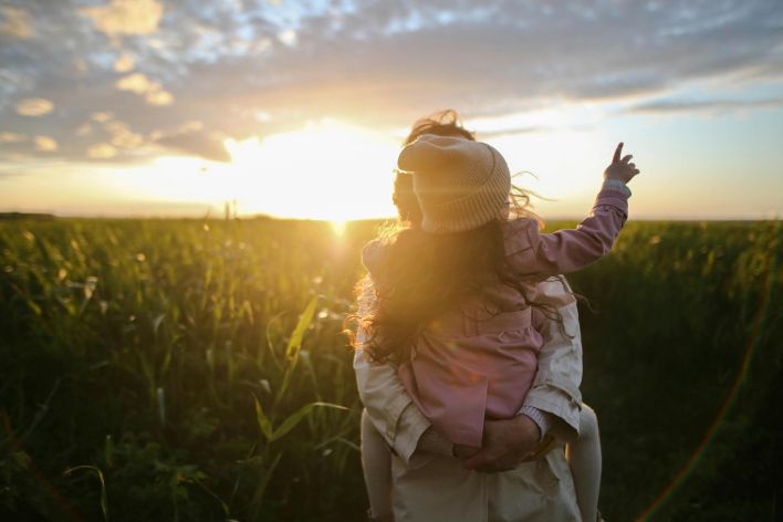 A mother and daughter embrace and point at the sunset in a grassy field.