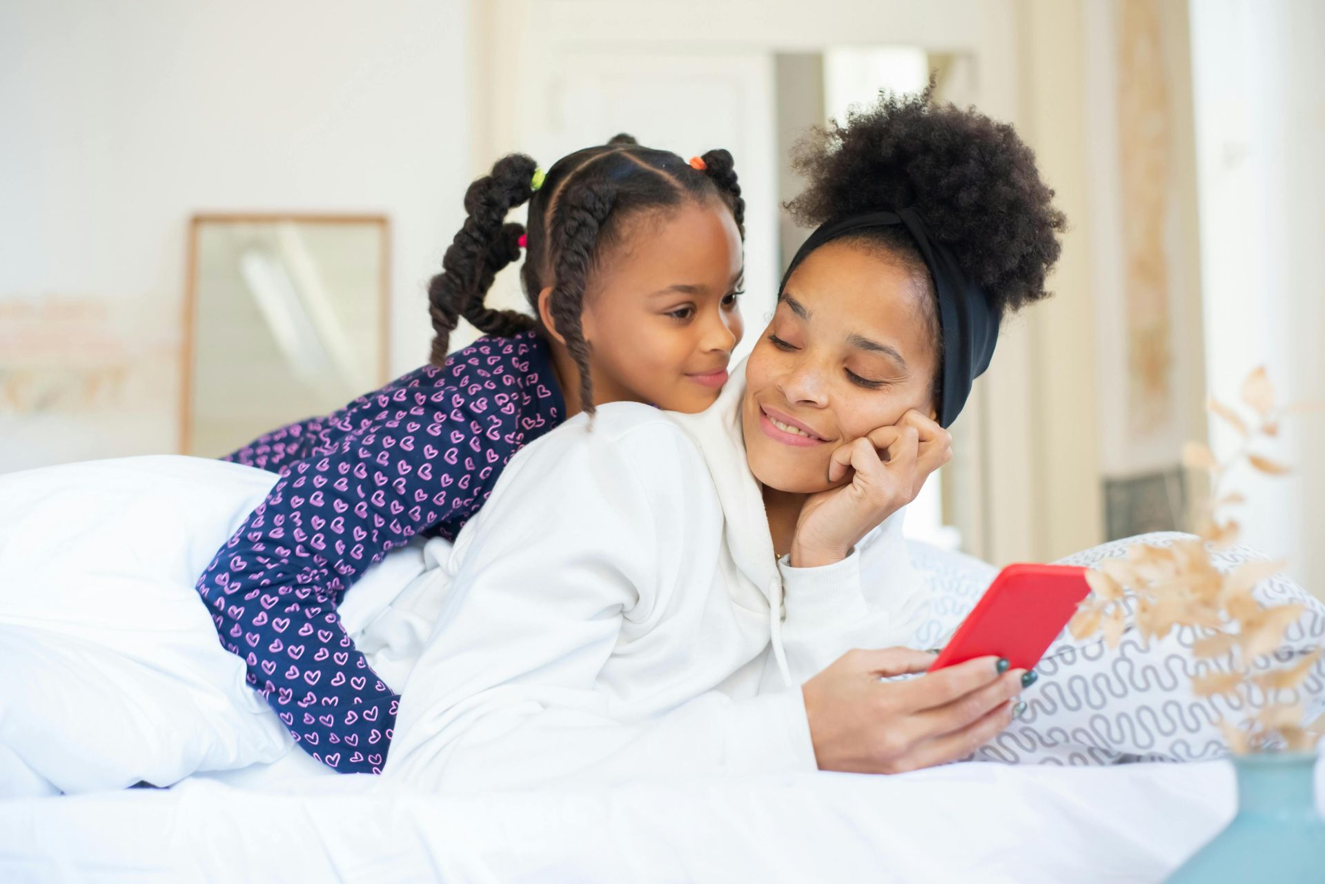 A heartwarming moment between a mother and daughter embracing while using a smartphone indoors.